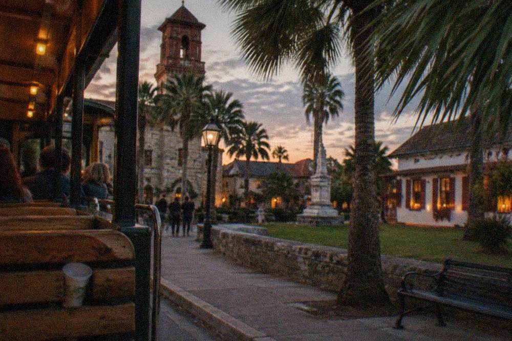 An open-sided trolley with wooden seats passing candle-lit windows of a historic street at dusk