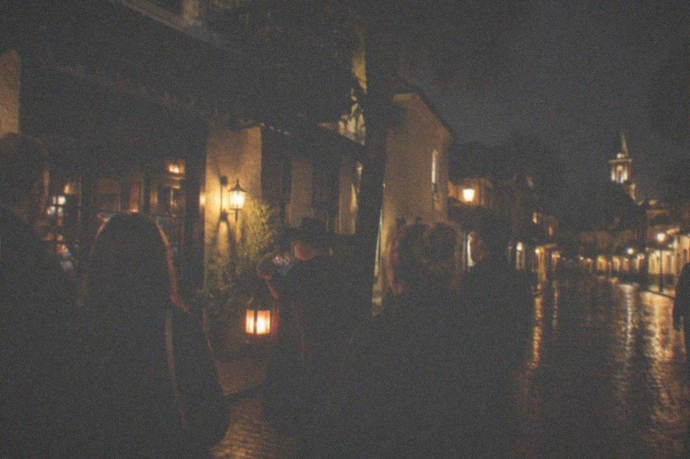 A lantern-lit historic street corner with a small crowd gathered around a guide at night